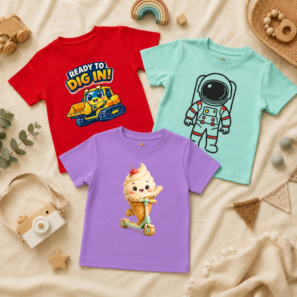 A flatlay of three kid's t-shirts arranged on a textured cream blanket alongside a wooden toy camera, a wooden rainbow, and natural woven decor. The shirts include a red t-shirt featuring a cartoon yellow bulldozer and the text "READY TO DIG IN!", a spearmint t-shirt displaying a stylized line-art graphic of an astronaut, and a lavender t-shirt showing a cheerful soft-serve ice cream cone riding a scooter.