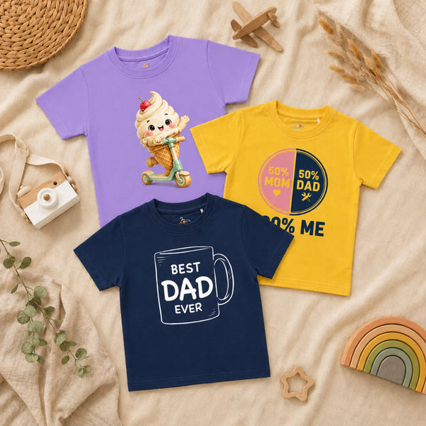 A flatlay of three kid's t-shirts arranged on a textured cream blanket alongside a woven mat, a wooden toy camera, a wooden toy airplane, a wooden rainbow, and dried foliage. The shirts include a lavender t-shirt showing a cheerful soft-serve ice cream cone riding a scooter, a sunshine yellow t-shirt with a split pink and navy circle graphic reading "50% MOM, 50% DAD" and "100% ME" below it, and a navy blue t-shirt displaying a white line drawing of a mug with the text "BEST DAD EVER".