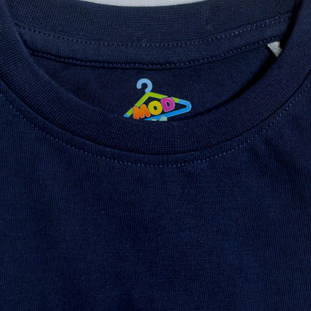 Close-up of a navy blue crewneck t-shirt showing the inner neck label with a colorful embroidered hanger logo that spells 'MOD 11' fabric-closeup